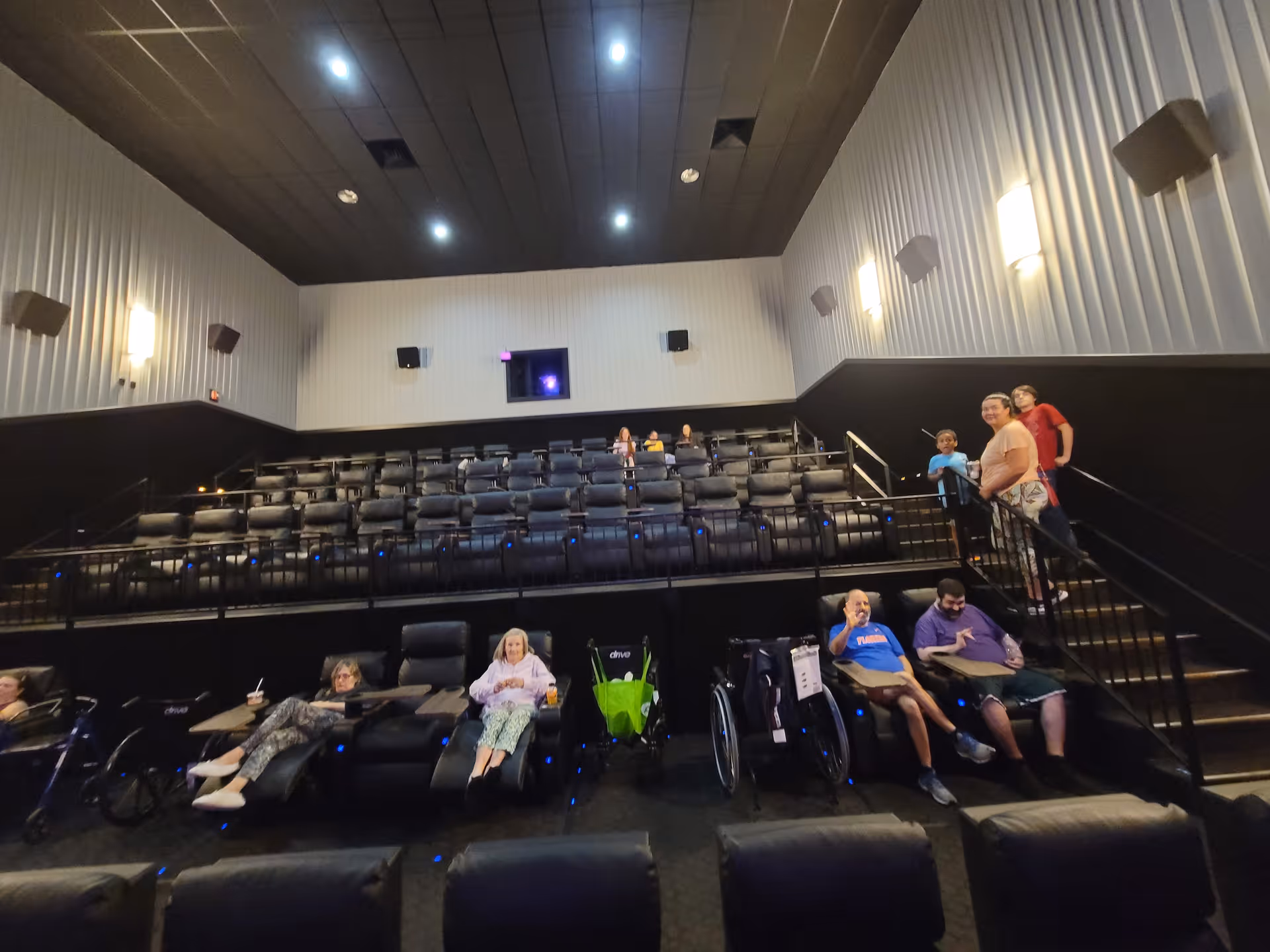Interior of a small movie theater with black reclining seats arranged in rows. Several people, including some in wheelchairs, are seated or standing on the stairs. The walls are light-colored with mounted lights, and the ceiling is dark with recessed lighting.