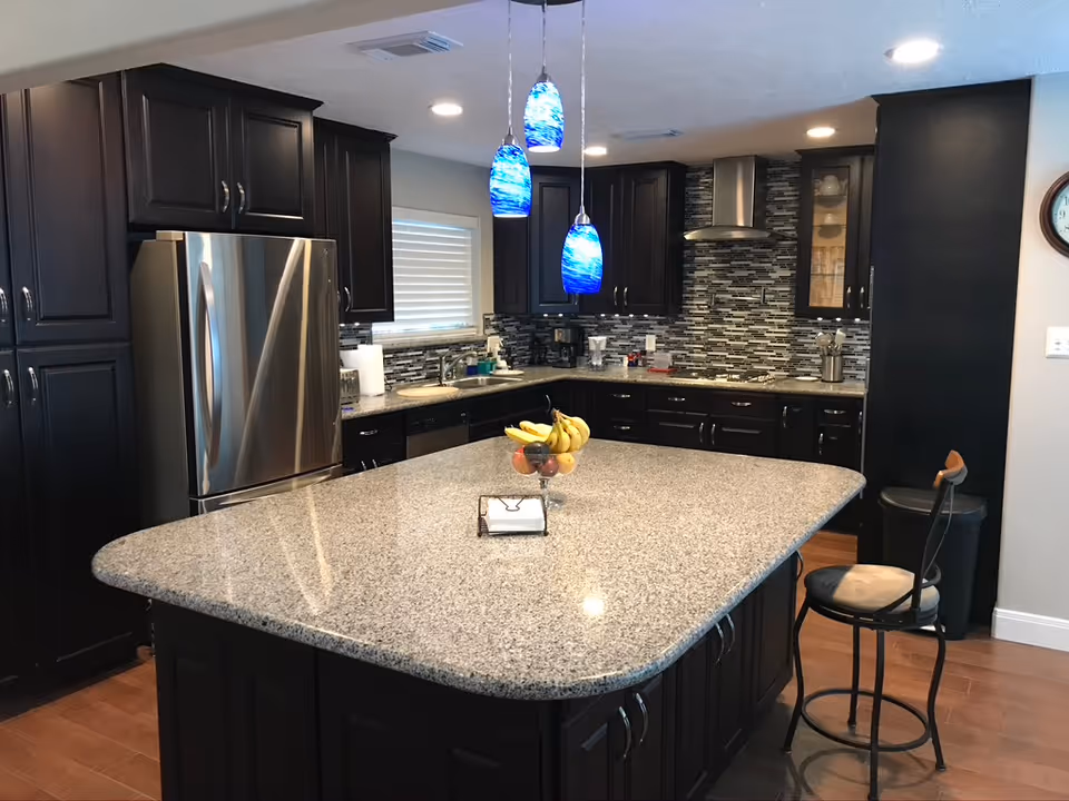 Modern kitchen with a large granite island, dark cabinets, stainless refrigerator, blue pendant lights and a bar stool.