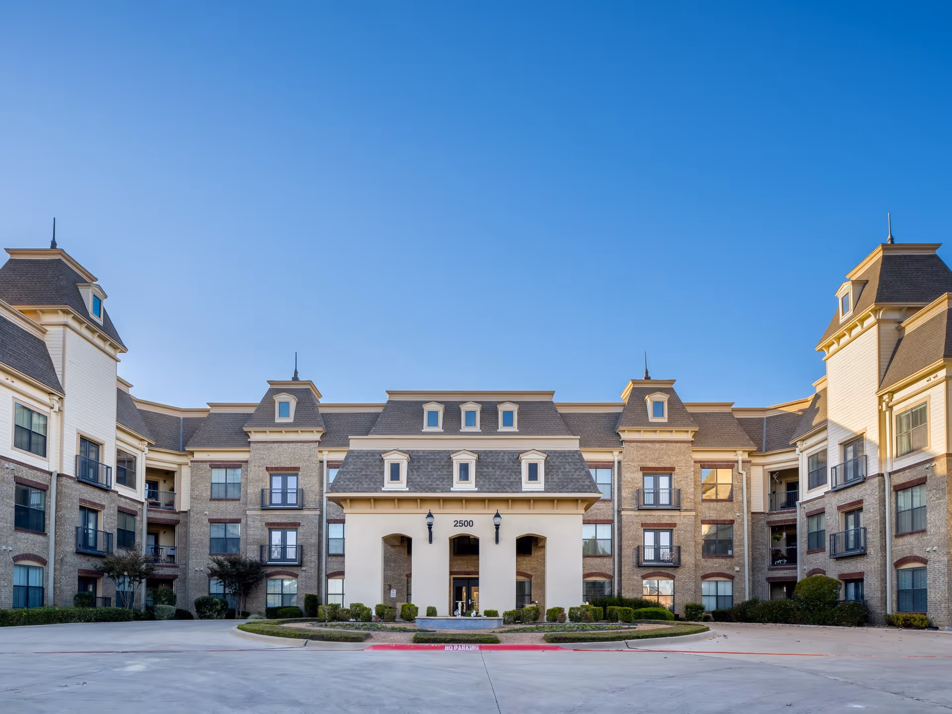 Front exterior view of a large, multi-story senior living facility building with a symmetrical design, multiple windows, and a central entrance under a clear blue sky.