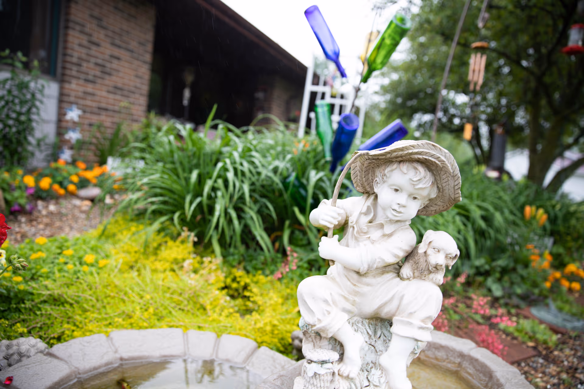 A garden scene featuring a white statue of a young boy wearing a hat and holding a fishing rod, with a small dog on his shoulder. The statue is placed near a small water feature surrounded by lush green plants and colorful flowers. In the background, there is a brick building and decorative blue and green glass bottles on stakes.