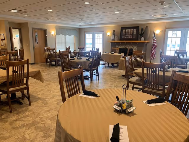 A spacious dining room with multiple round and rectangular tables covered with beige tablecloths, each set with black napkins and small condiment holders. The room has wooden chairs, a tiled floor, large windows letting in natural light, a fireplace with decorative items above it, and an American flag in the corner.
