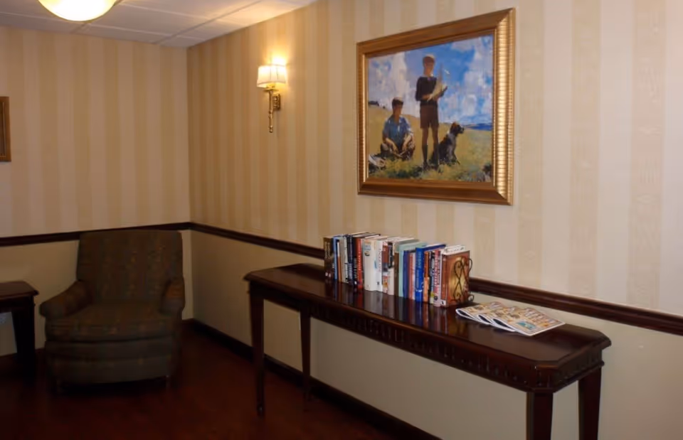 A cozy corner of a room with a patterned armchair, a wooden side table, and a long wooden console table holding a row of books and some magazines. Above the console table hangs a framed painting depicting two boys and a dog in a grassy field under a blue sky with clouds. The walls have beige vertical striped wallpaper with a chair rail and a wall sconce light fixture.