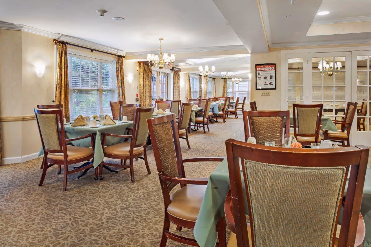 A dining room in a senior living facility with multiple tables covered in green tablecloths, each set with napkins and cups. The room has large windows with patterned curtains, carpeted floors, and chandeliers hanging from the ceiling. Wooden chairs with cushioned seats surround the tables.