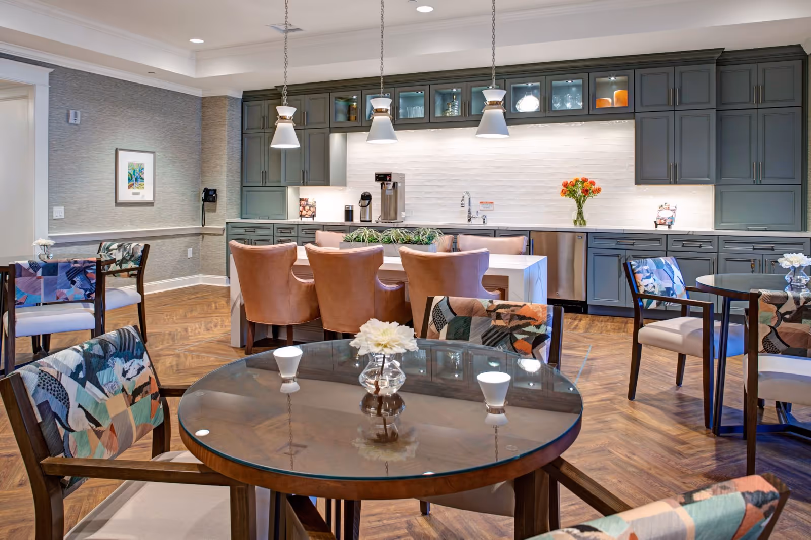 Bright communal dining area with round glass tables, patterned chairs, a central island with leather stools, and a kitchenette with gray cabinets.