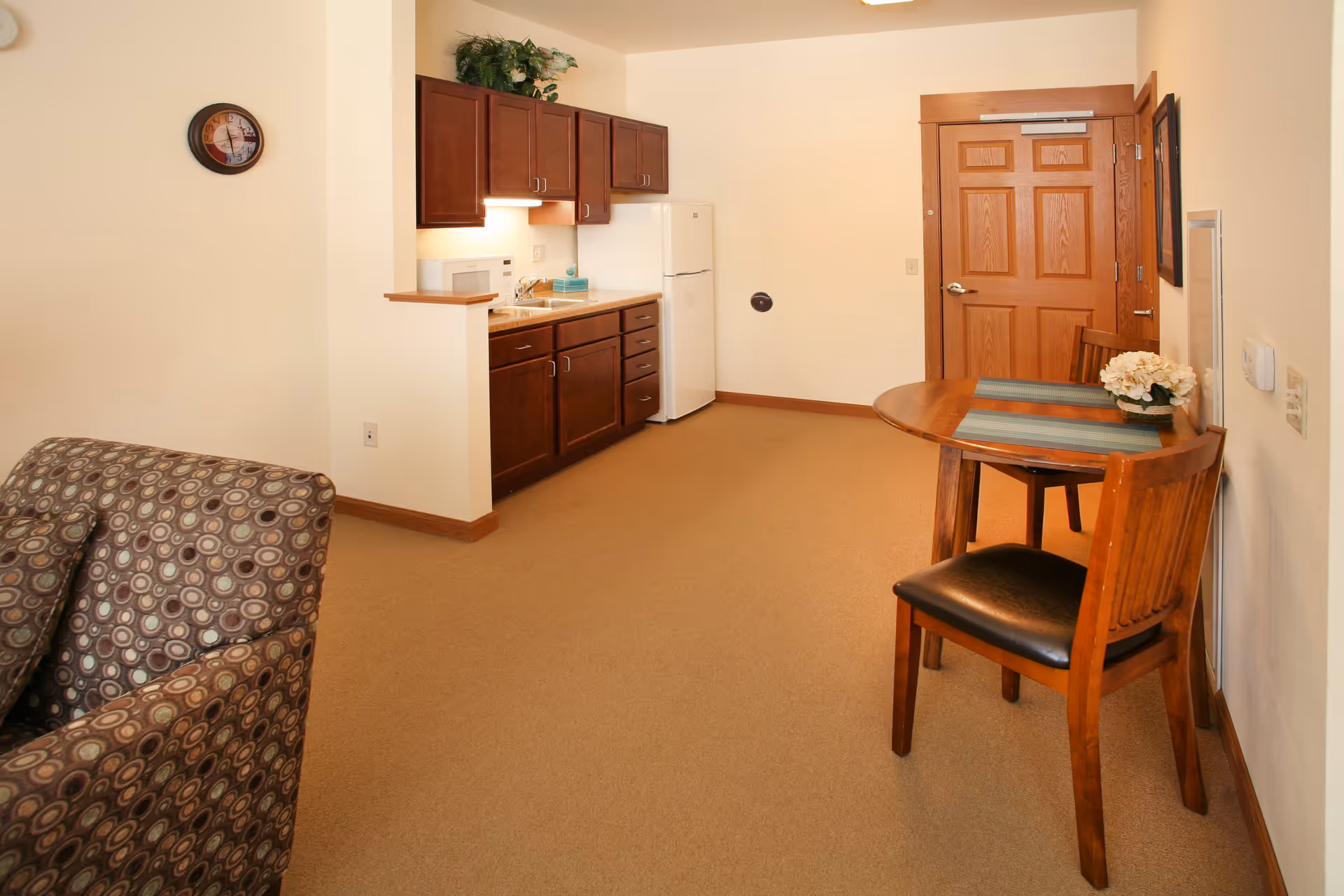 Interior view of a small apartment or living space featuring a kitchenette with dark wood cabinets, a white refrigerator, and a microwave. There is a round wooden table with two chairs, one of which has a black seat cushion, and a small flower arrangement on the table. A patterned armchair is partially visible on the left side. The walls are light-colored, and there is a wooden door in the background.