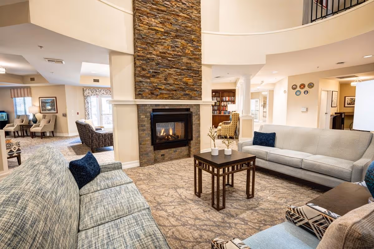 A cozy living room area in a senior living facility featuring two sofas with blue cushions, a stone fireplace with a fire burning, a wooden coffee table with decorative vases, patterned carpet, and additional seating and bookshelves in the background.