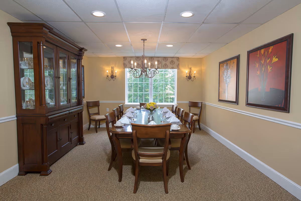 A formal dining room with a long wooden table set for a meal, chairs, a china cabinet, chandelier, and a window showing greenery.
