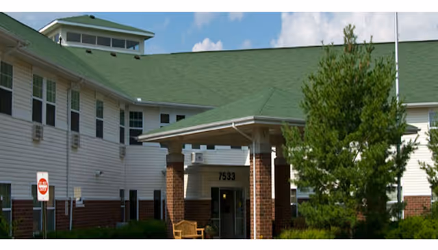 Exterior view of a senior living facility building with a green roof, white siding, and brick accents. The entrance has a covered porch supported by brick columns, with the number 7533 displayed above the door. There is a tree and some greenery near the entrance, and a stop sign is visible on the left side.