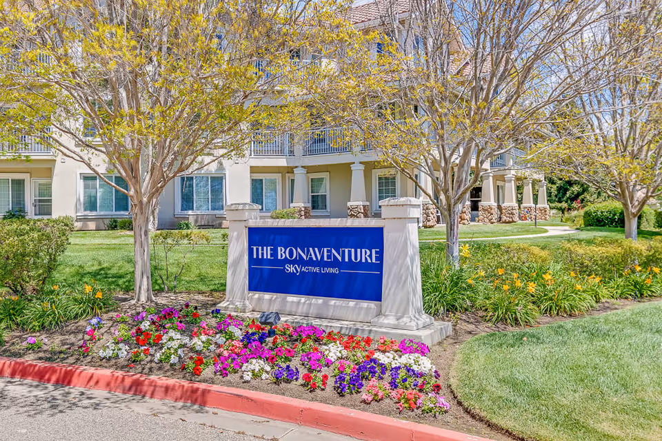 Outdoor view of a senior living facility with a landscaped garden featuring colorful flowers and trees. A blue sign in the foreground reads 'THE BONAVENTURE SKY ACTIVE LIVING' with a multi-story building in the background.