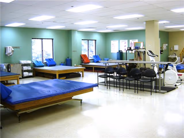 A spacious rehabilitation room with multiple padded therapy tables, chairs, exercise equipment, and parallel bars. The room has light green walls, large windows letting in natural light, and a clean, bright atmosphere.