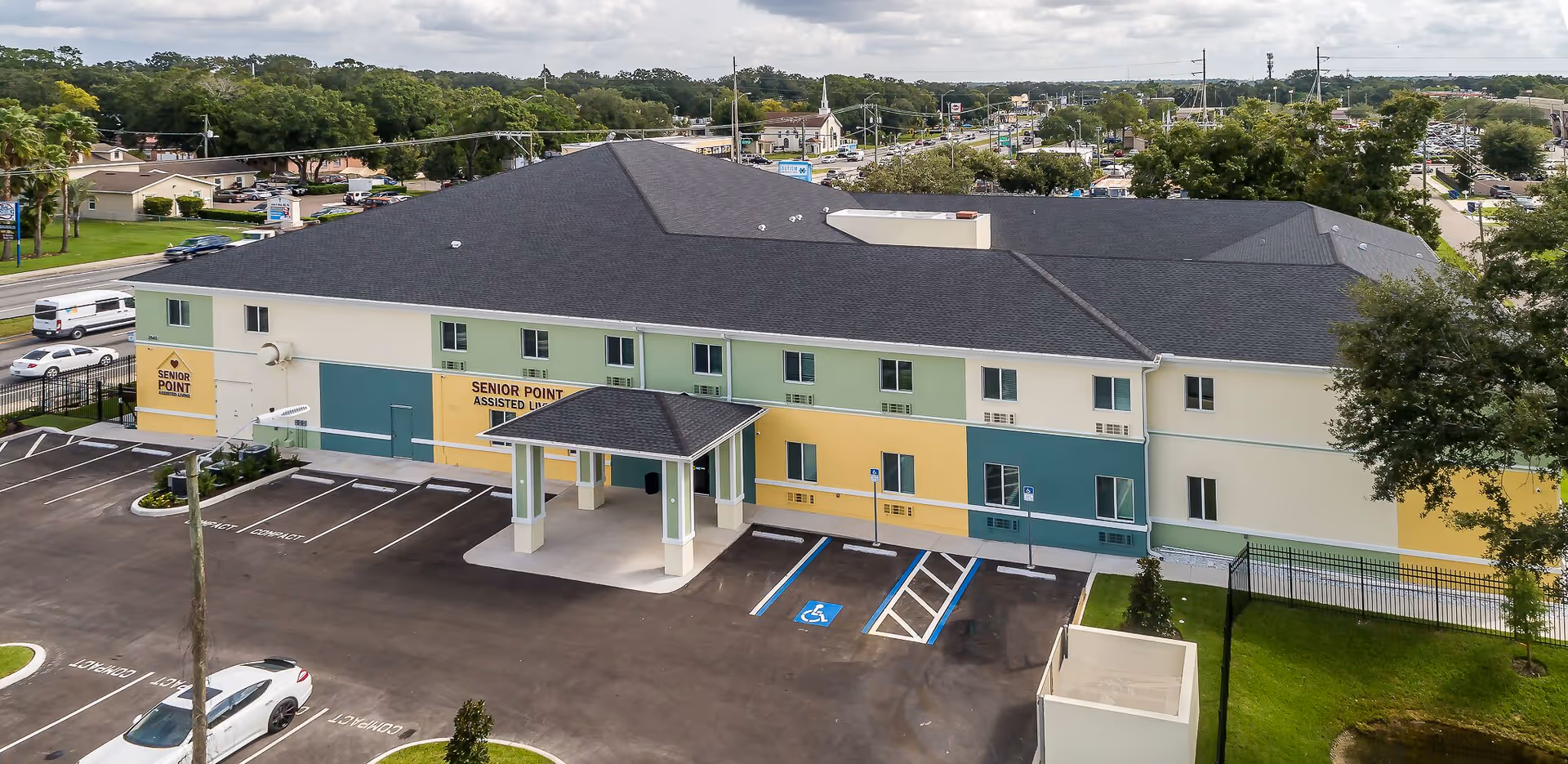 Aerial view of Senior Point Assisted Living facility showing a two-story building with a multi-colored exterior in shades of green, yellow, and cream. The building has a covered entrance and several windows. There is a parking lot in front with marked compact and handicapped parking spaces. Surrounding the building are trees, grass, and a busy street with cars and other buildings in the background under a cloudy sky.