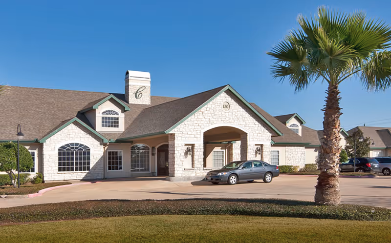 Exterior view of Carriage Inn Lake Jackson building with stone facade, arched entrance, and a parked car in front under a clear blue sky. A palm tree and trimmed bushes are visible in the landscaped area.