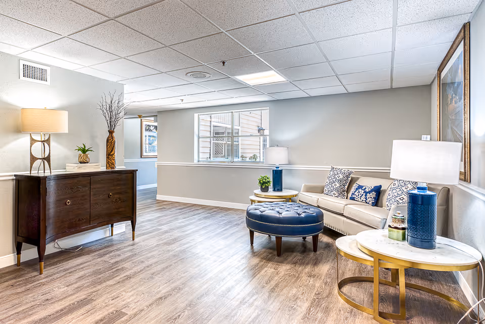 A bright and modern living room area with a beige sofa adorned with patterned cushions, a round blue ottoman, two round side tables with white marble tops and gold frames, each holding a blue lamp. There is a dark wooden sideboard with a lamp, a small plant, and a decorative vase with branches. The room has light gray walls, wood flooring, and a large window letting in natural light.