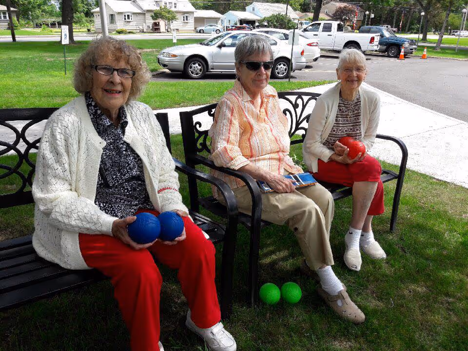 Three elderly women sitting on outdoor benches on a grassy area. Two of the women are holding bocce balls, one with two blue balls and the other with a red ball. The third woman is holding a tablet. There are cars and houses visible in the background.