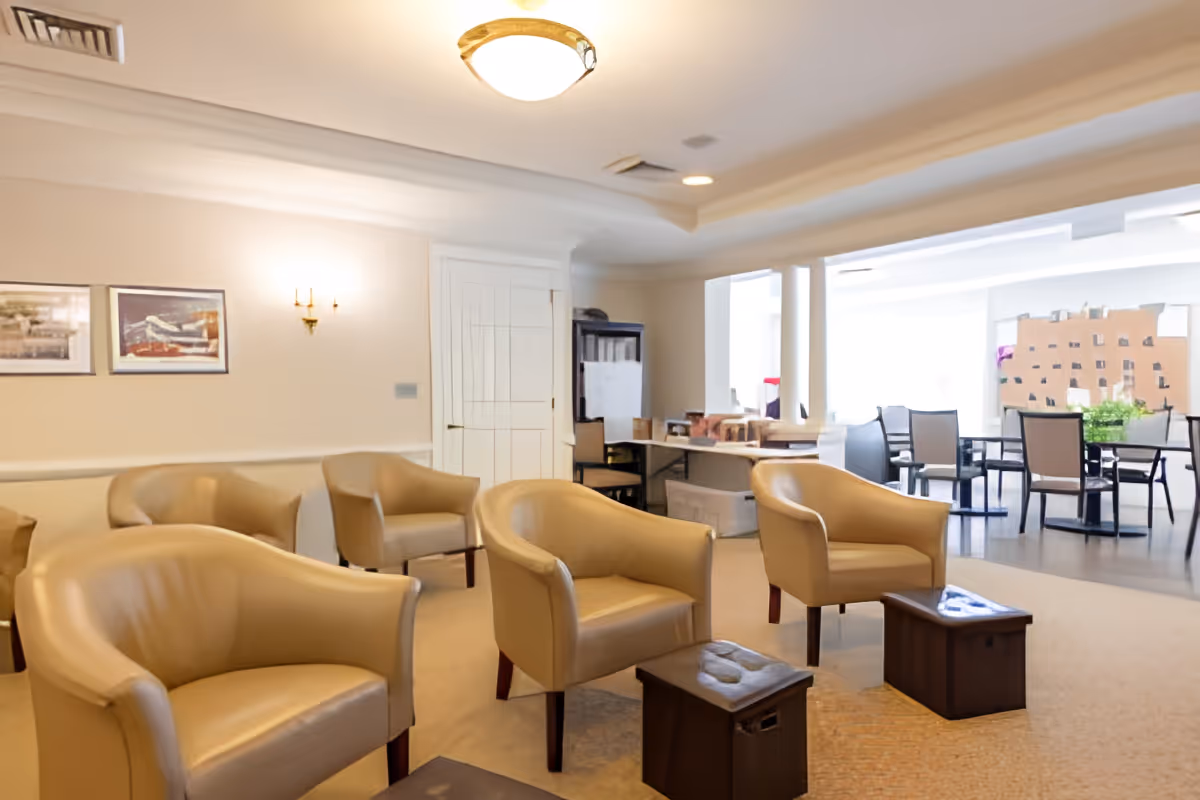 Bright communal lounge in a senior living facility with beige armchairs, small ottomans, and a dining area visible through an open doorway.