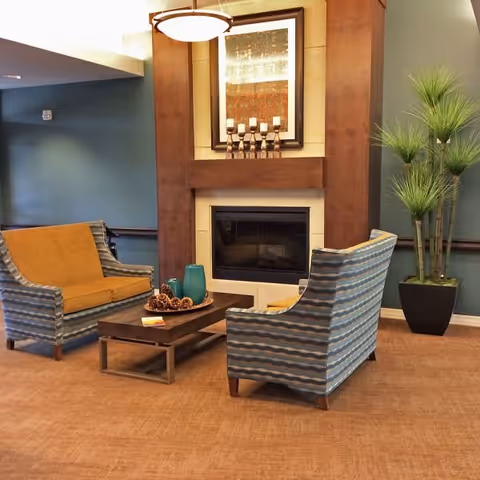 A cozy seating area in a senior living facility featuring two armchairs with patterned upholstery and mustard yellow cushions, a wooden coffee table with decorative items, a modern fireplace with a wooden mantel, a framed artwork above the fireplace, and a tall potted plant in the corner.