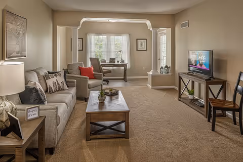 Cozy neutral-toned living room with a sofa and armchairs around a wooden coffee table, a TV on a console, and a desk by a window.