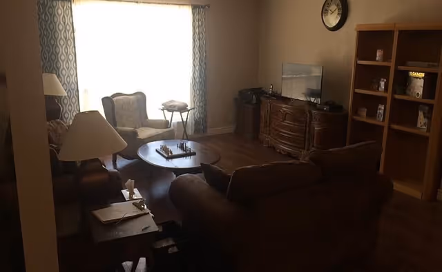 A cozy living room with a brown sofa and armchair, a wooden coffee table with a chessboard on it, a TV on a wooden cabinet, a bookshelf with various items, and a window with patterned curtains letting in natural light.