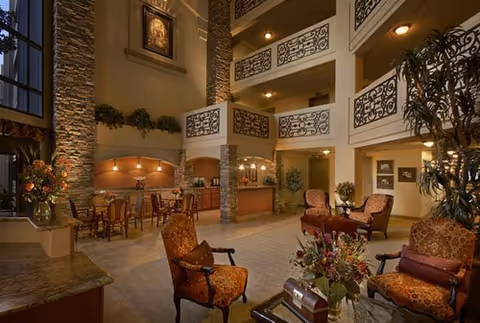 Spacious and warmly lit senior living facility common area with high ceilings, stone pillars, decorative railings on upper balconies, and comfortable upholstered chairs arranged around a glass coffee table with a floral centerpiece. A dining area with tables and chairs is visible in the background near a kitchen or serving counter.
