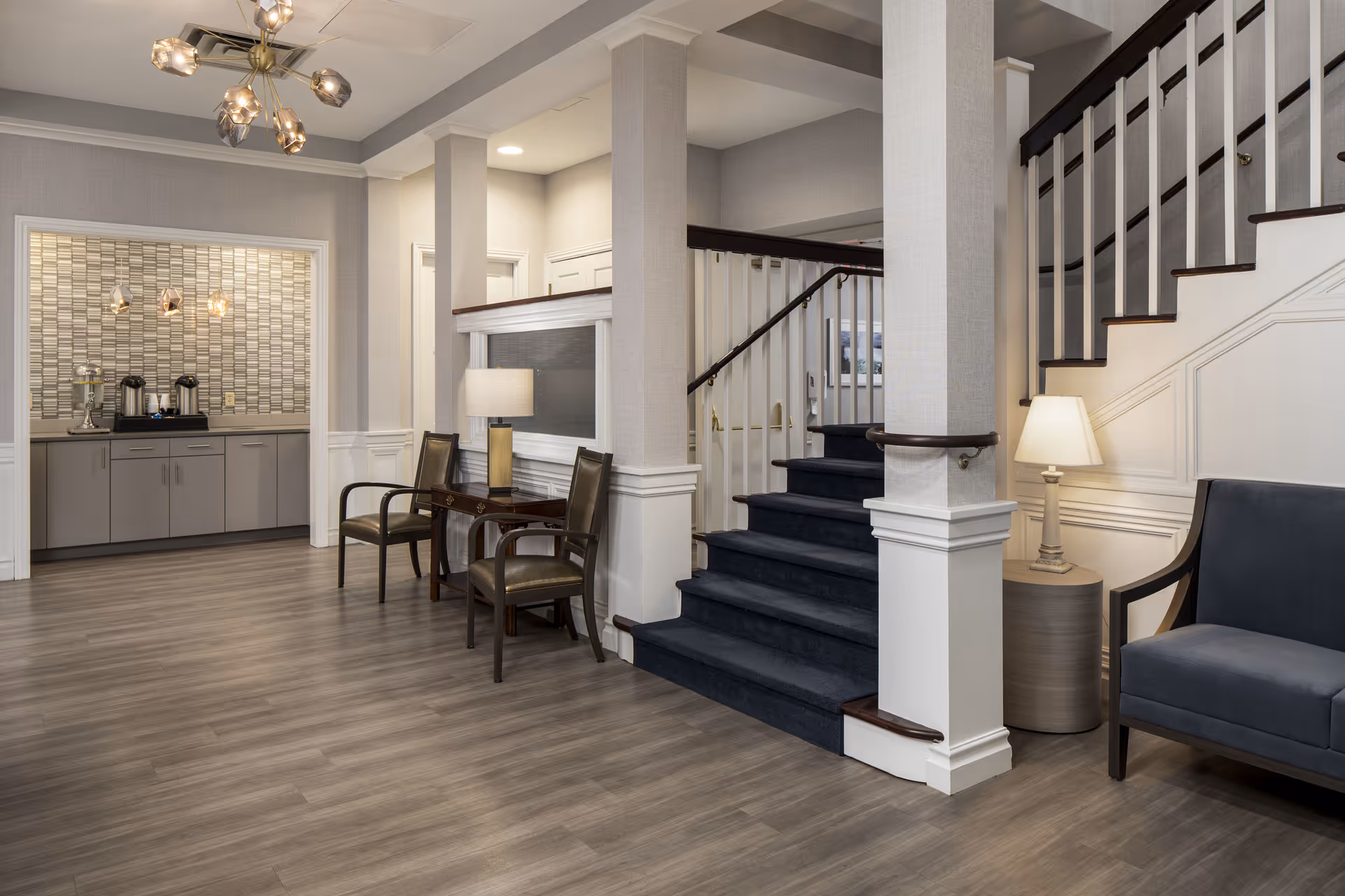 Bright furnished lobby with seating, a carpeted staircase, and a coffee station in the background.