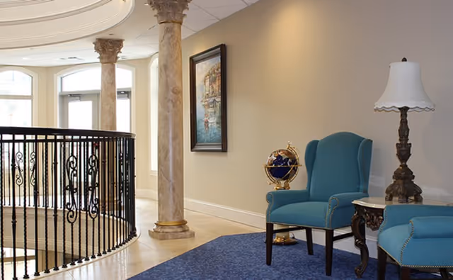 An elegant senior living facility interior with teal armchairs, a side table and lamp, decorative globe, marble columns, and a wrought-iron railing around a staircase.