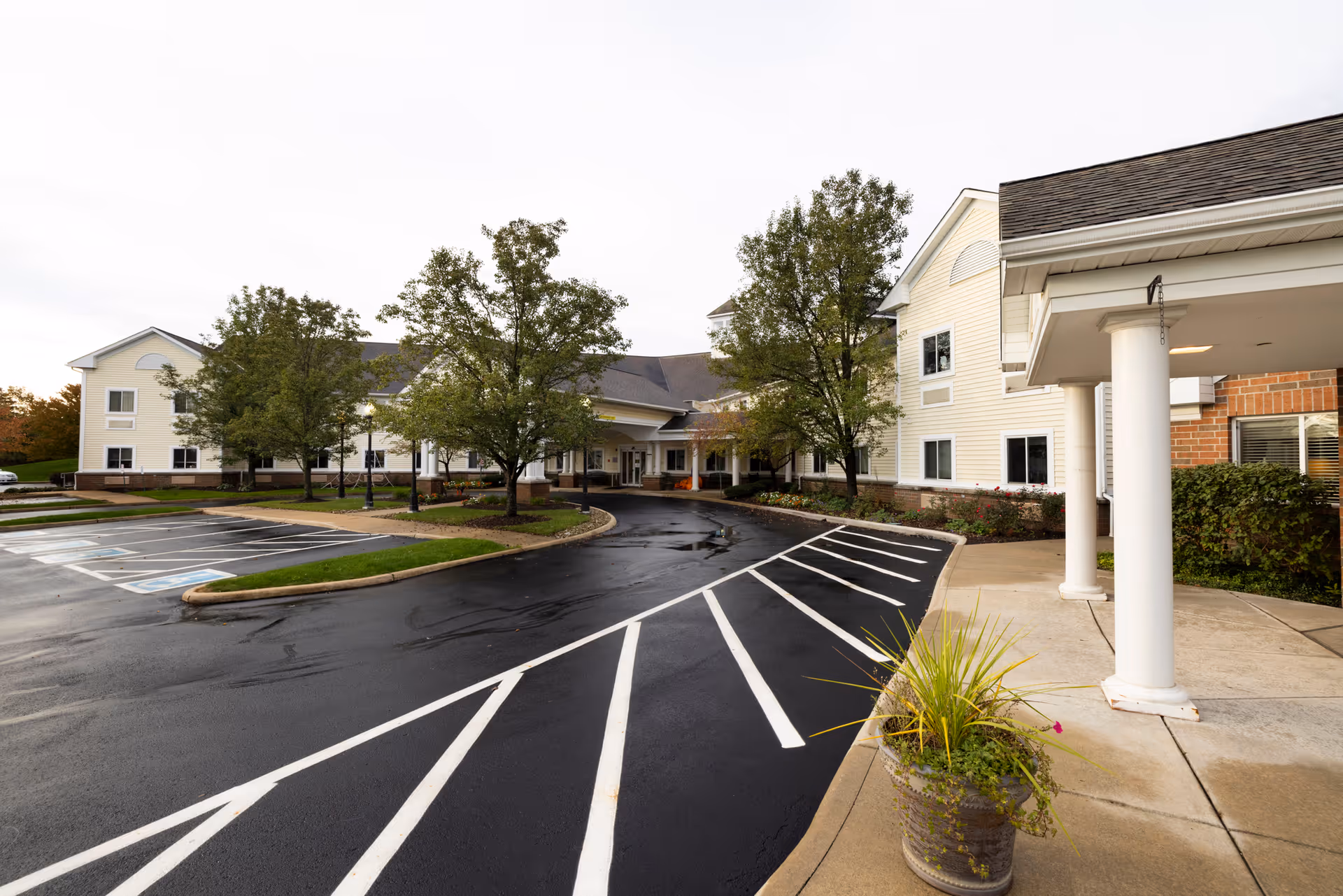 Exterior view of Canterbury Senior Care Facility showing a large, light-colored building with multiple windows, a covered entrance supported by white columns, a paved driveway with marked parking spaces including handicapped spots, and several trees and landscaped areas.