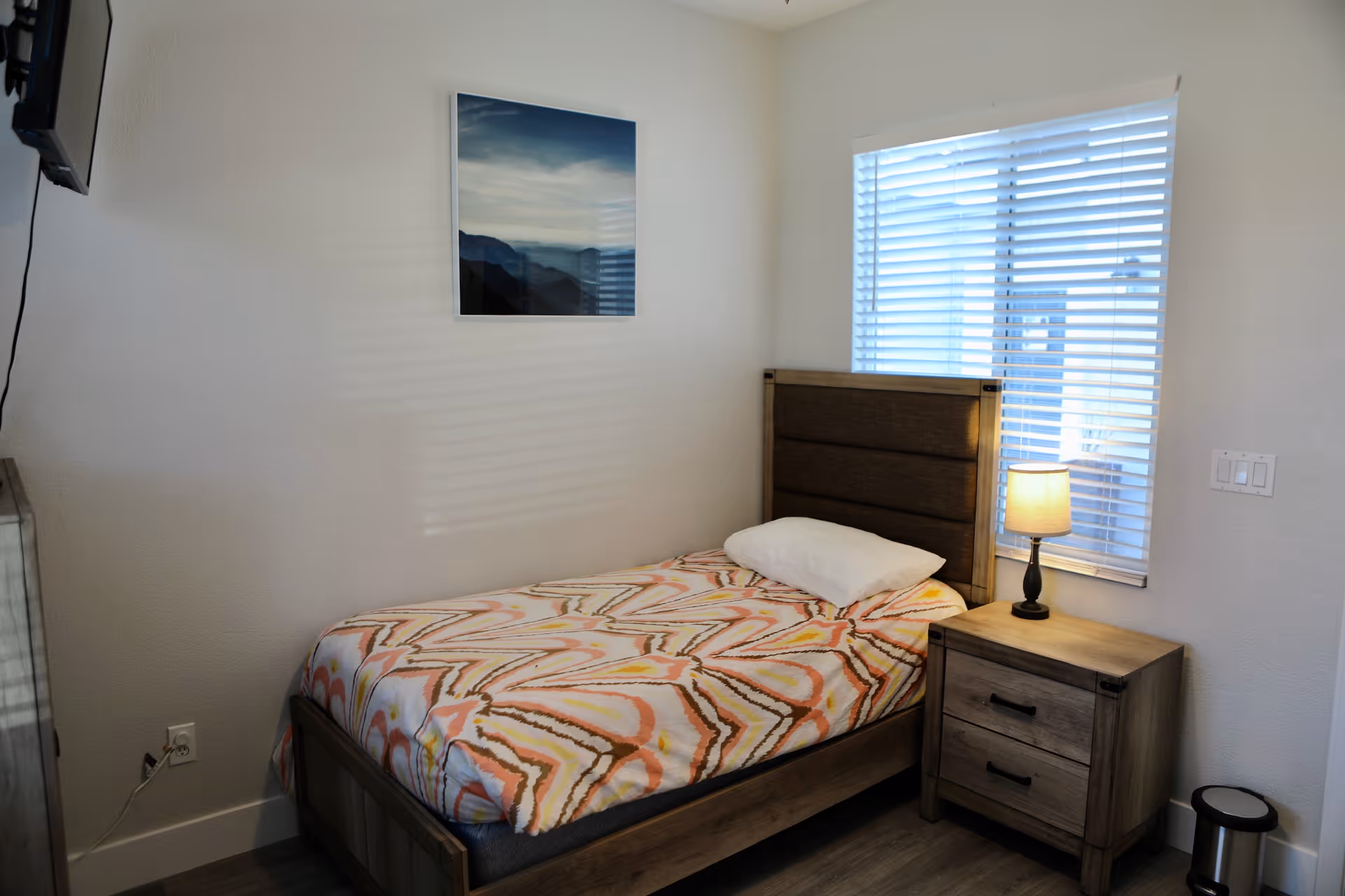 Simple single bedroom with a patterned bed, wooden headboard and nightstand with a lamp beneath a window with blinds.