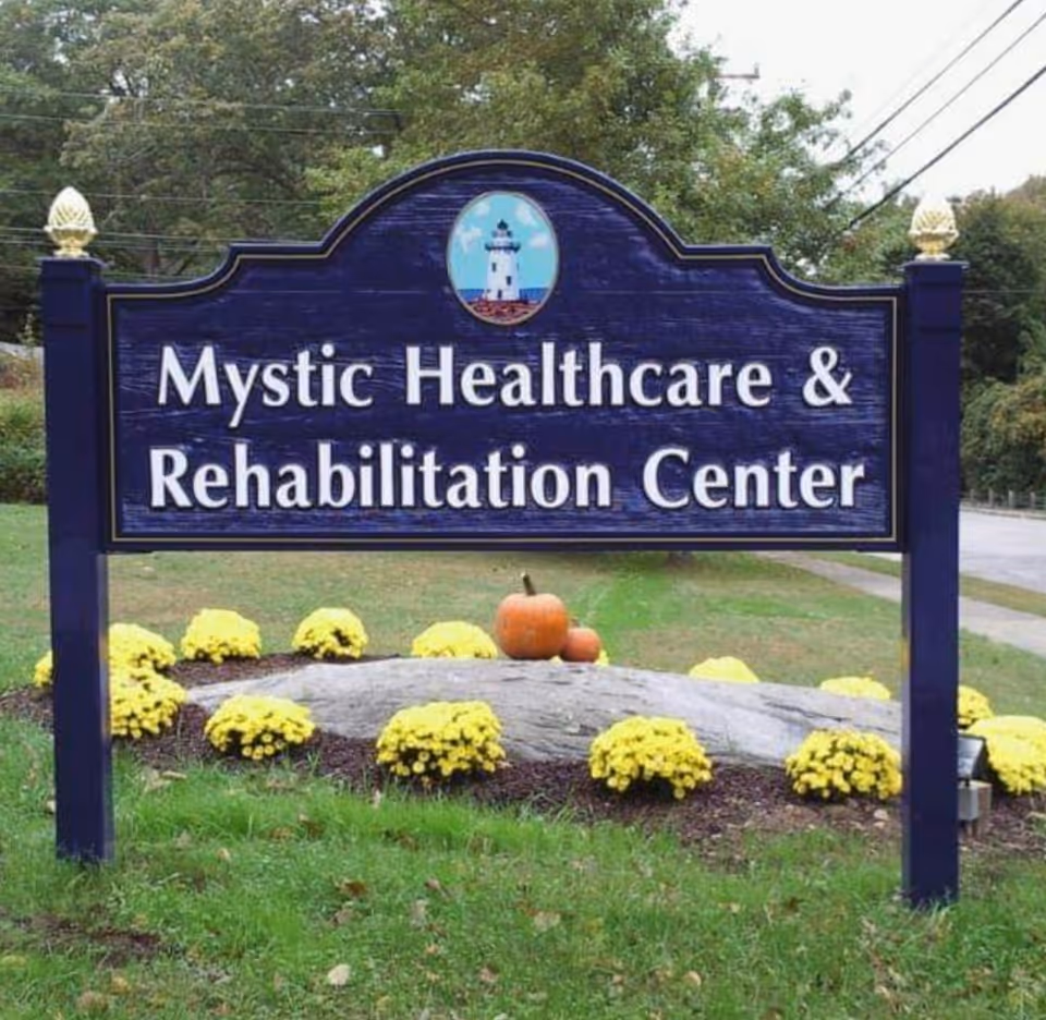 Outdoor sign for Mystic Healthcare & Rehabilitation Center, surrounded by yellow flowers and two pumpkins, with trees and a road in the background.