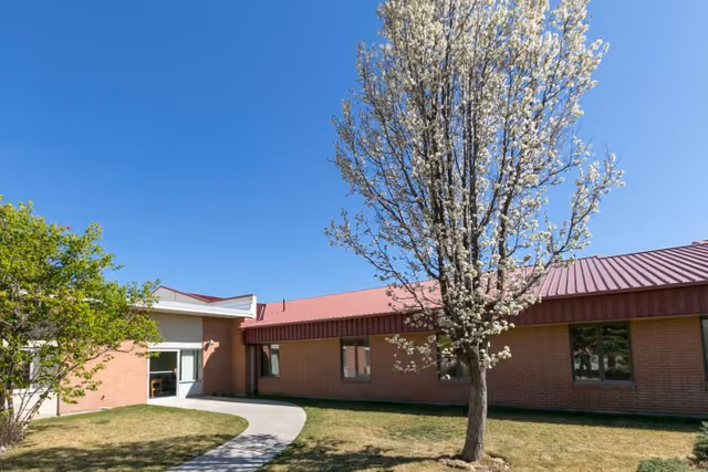Exterior view of Gateway Transitional Care Center showing a single-story brick building with a red metal roof, a blossoming tree in the foreground, a curved concrete walkway, and a clear blue sky.