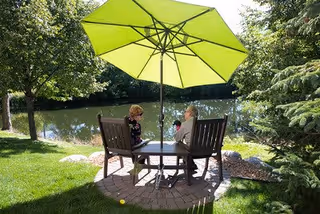 Two elderly women sitting at a round outdoor table with a large green umbrella, surrounded by trees and greenery, next to a calm body of water.