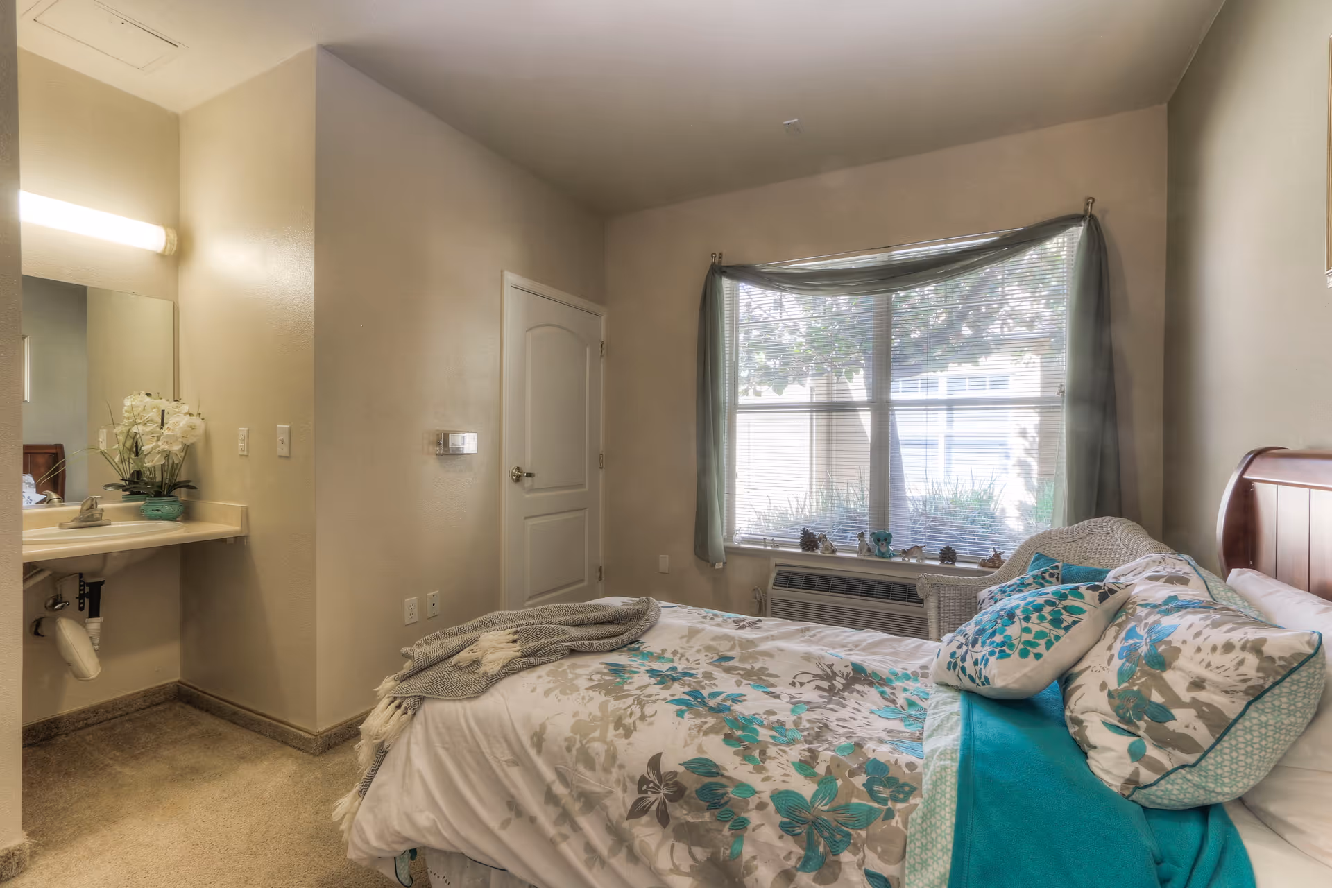 A cozy bedroom with a bed covered in floral patterned bedding in shades of teal, gray, and white. There are multiple pillows and a teal blanket on the bed. To the left, there is a small sink with a mirror above it and a vase with white flowers. A window with blinds and sheer teal curtains lets in natural light, and a white wicker chair is placed near the window. The walls are painted beige and the carpet is light brown.