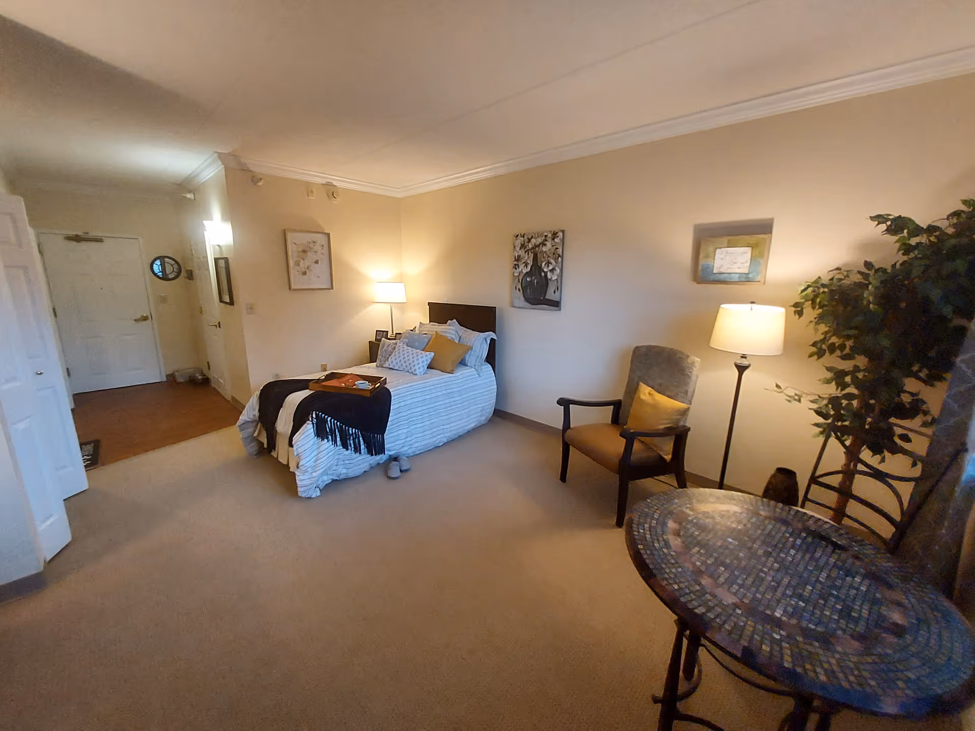 A cozy senior living bedroom with a single bed made with striped bedding and multiple pillows. A tray with items is placed on the bed. There is a cushioned armchair with a yellow pillow next to a floor lamp and a potted plant. A round mosaic table with two chairs is in the foreground. The room has beige walls with framed artwork and a carpeted floor. The entrance door and hallway are visible in the background.
