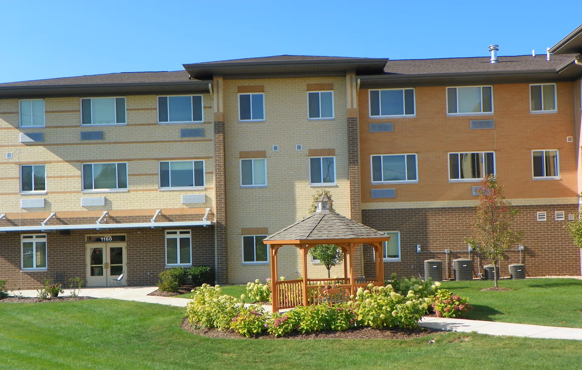 Exterior view of a multi-story senior living facility building with a well-maintained lawn and a wooden gazebo surrounded by shrubs and flowers in the foreground under a clear blue sky.