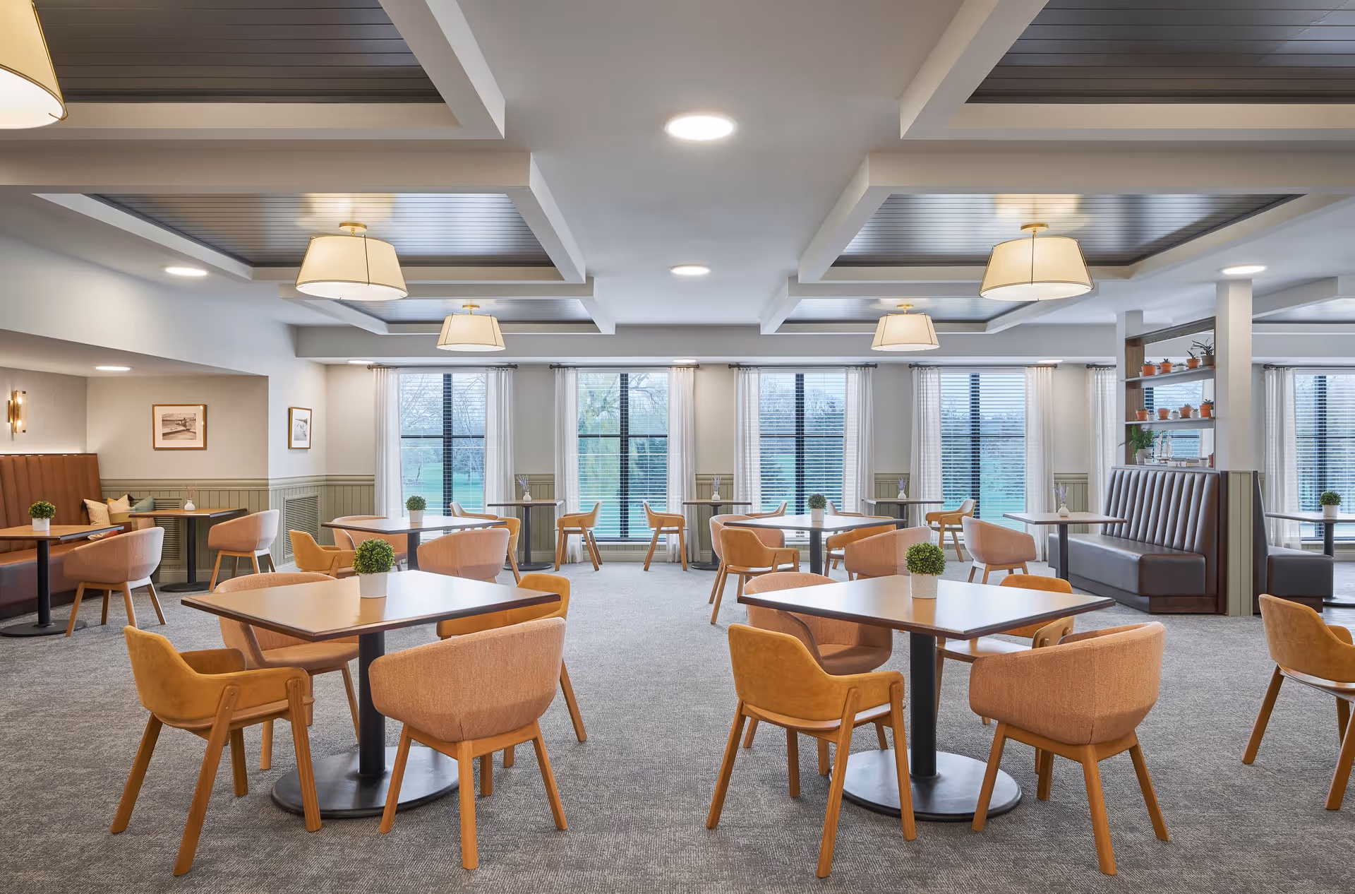A bright and spacious dining room with multiple square tables, each surrounded by four cushioned chairs in a warm beige color. The room features large windows with white curtains allowing natural light to fill the space. The ceiling has recessed lighting and decorative hanging lamps. There are also booth seating areas along the walls with framed artwork and small potted plants on each table.