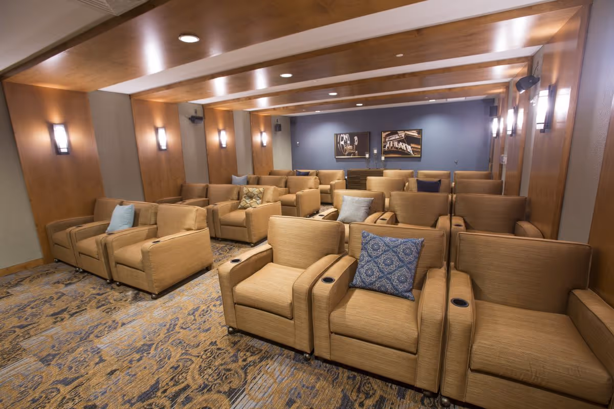 A cozy theater room with multiple rows of tan upholstered armchairs, each with cup holders. The room has wood paneling on the walls and ceiling beams, soft wall lights, and framed artwork on the far wall. The carpet has a patterned design in shades of blue and beige.