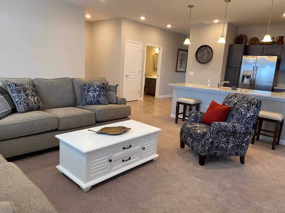 A cozy living room area featuring a gray sofa with patterned cushions, a white coffee table with a decorative leaf tray, and a patterned armchair with a red pillow. In the background, there is a kitchen with a granite countertop, bar stools, pendant lights, and stainless steel refrigerator. The walls are painted light gray and the floor is carpeted.