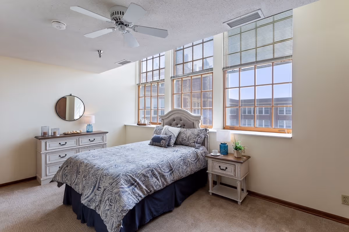A bright bedroom with a large bed covered in a patterned blue and gray comforter and multiple pillows. The room features three large windows with wooden frames and blinds, letting in natural light. There is a white ceiling fan above, a white dresser with a round mirror and a lamp on the left side, and a matching nightstand with a lamp, a small plant, and a cup on the right side. The walls are painted light beige and the floor is carpeted.