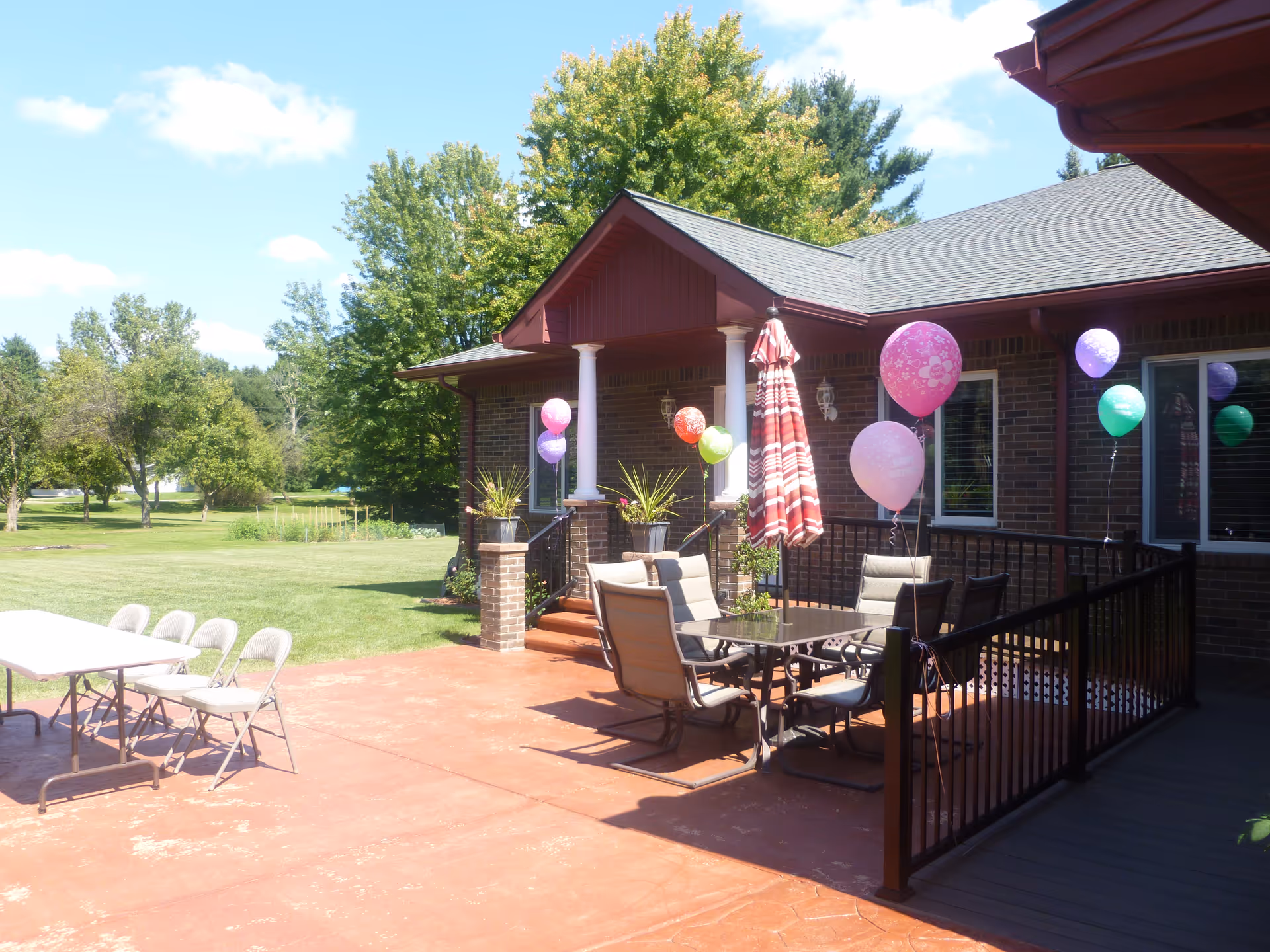 Outdoor patio area at a senior living facility with a table and chairs, decorated with colorful balloons and an umbrella. The patio is adjacent to a brick building with white columns and surrounded by green trees and grass under a partly cloudy sky.