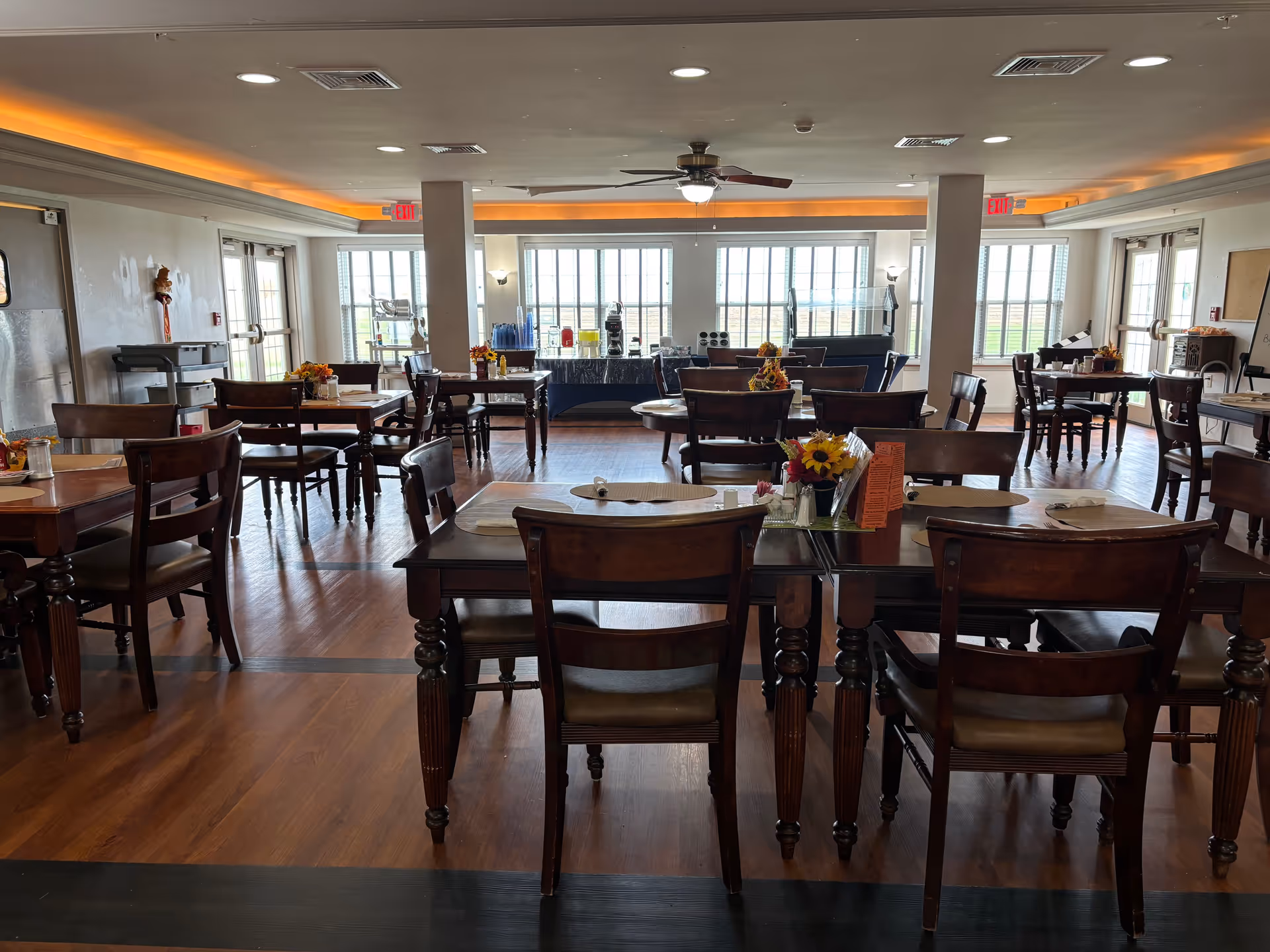 A spacious dining room with multiple wooden tables and chairs arranged neatly. Each table has placemats and small flower arrangements. Large windows at the far end allow natural light to fill the room. The ceiling has recessed lighting with a warm glow and a ceiling fan in the center. There are exit signs above the doors on both sides of the room.
