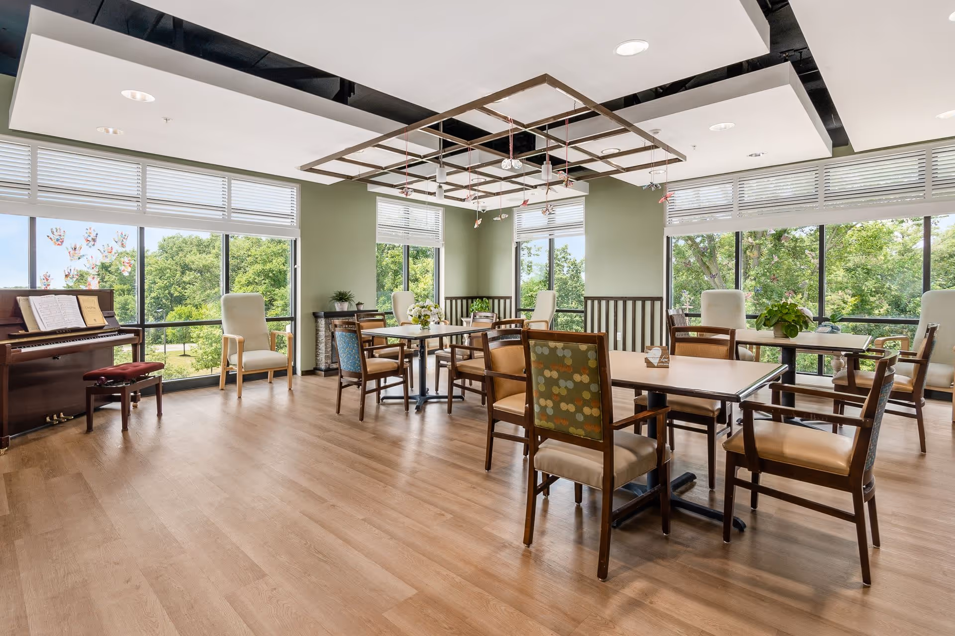 A bright and spacious room with large windows showing green trees outside. The room has several tables with chairs arranged around them, some with patterned upholstery. There is a piano with sheet music and a stool on the left side. The ceiling features a decorative grid with hanging lights. The floor is wooden, and the walls are painted light green.