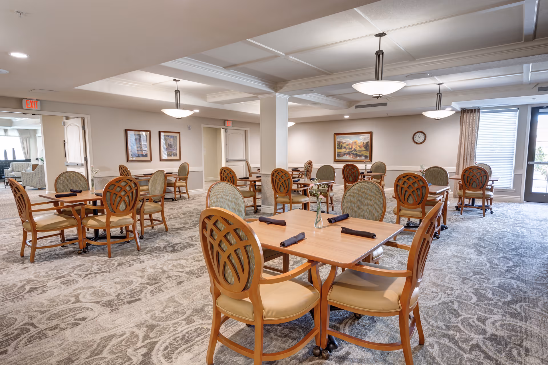 A bright senior living dining room with multiple wooden tables and chairs arranged on a patterned carpet, each place set with a folded napkin and small centerpiece.