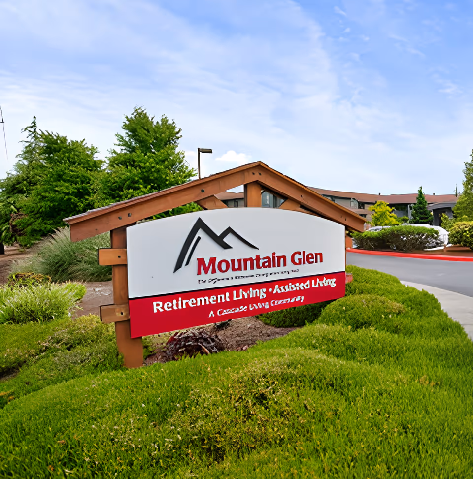 A wooden entrance sign for Mountain Glen Retirement and Assisted Living sits among green landscaping with the facility building and blue sky behind it.