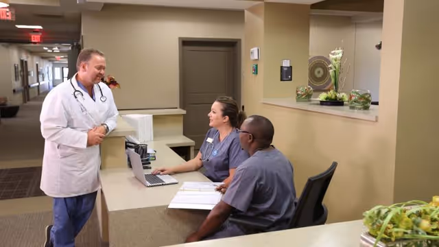 A doctor in a white coat is standing and talking to two healthcare workers seated at a reception desk in a medical facility. One healthcare worker is using a laptop while the other is looking at a document. The area has beige walls and a hallway in the background.