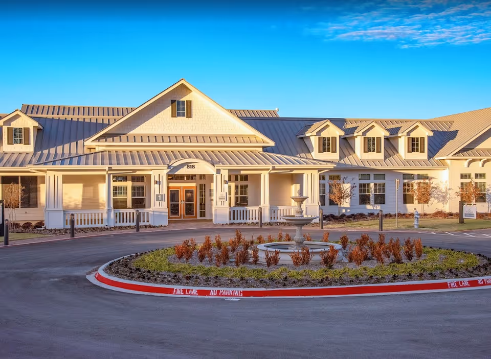 Front exterior view of a large, single-story building with a metal roof and multiple dormer windows. The entrance has double doors under a covered porch with white columns. In front of the building is a circular driveway with a landscaped island featuring a tiered fountain and small shrubs. The curb is painted red with 'FIRE LANE NO PARKING' markings.