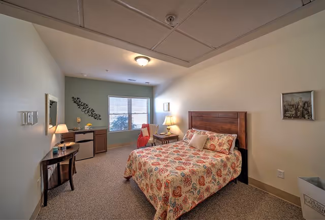 A cozy senior living bedroom with a double bed covered in a floral patterned quilt. The room features a wooden headboard, a bedside table with a lamp, a chair with a red throw blanket, a small desk with a mini fridge underneath, and a window with blinds letting in natural light. The walls are painted in light colors with a decorative wall decal and framed pictures.