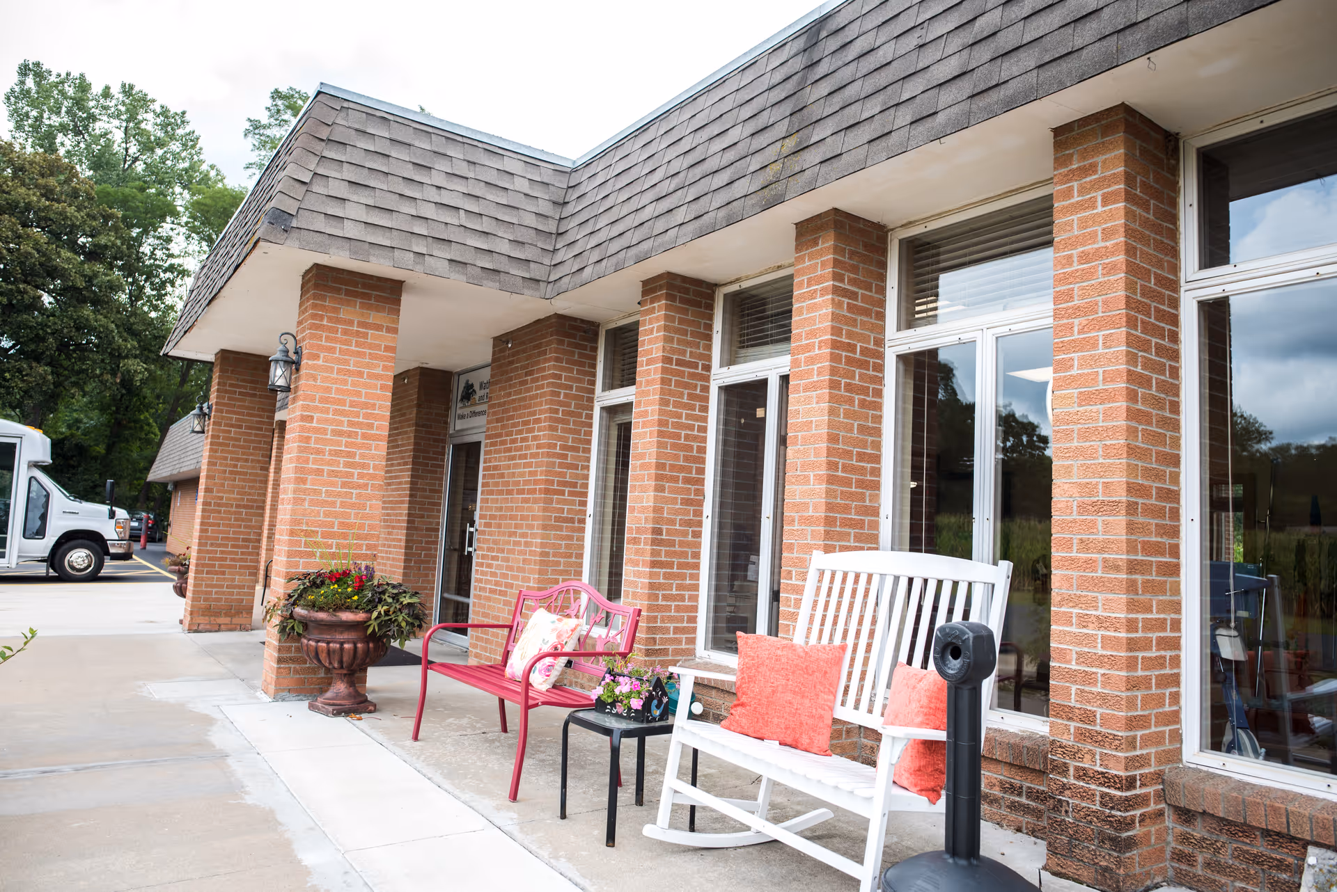 Exterior view of a brick building with large windows and a covered walkway. There are two benches, one white rocking bench with red cushions and one red metal bench with a floral cushion, along with a small black table holding a flower pot. A large planter with flowers is placed near the entrance door. Trees and a white vehicle are visible in the background.