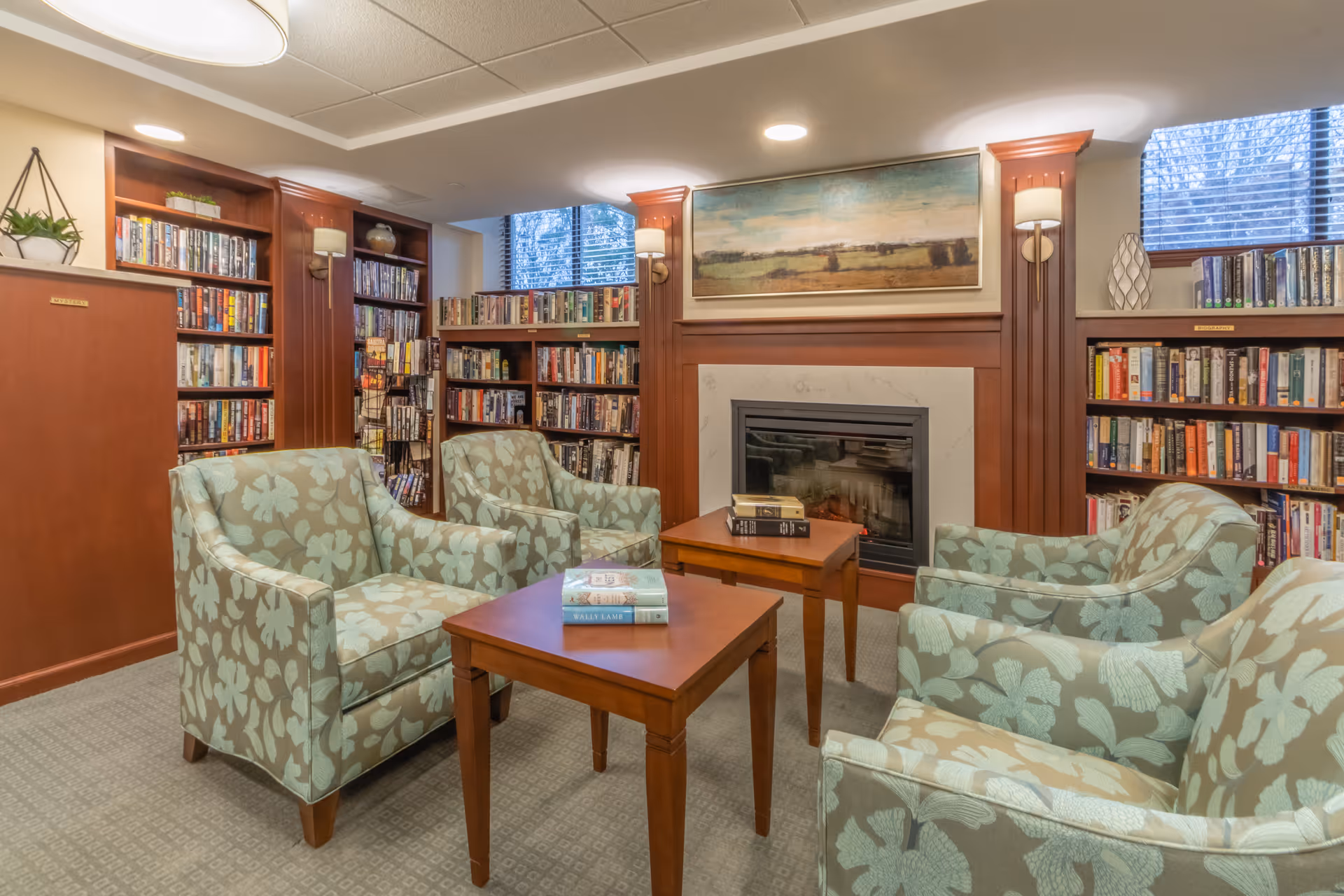 A cozy library room with four floral-patterned armchairs arranged around two wooden tables. The room features built-in wooden bookshelves filled with books, a fireplace with a marble surround, and a landscape painting above it. Two windows with blinds allow natural light into the space.