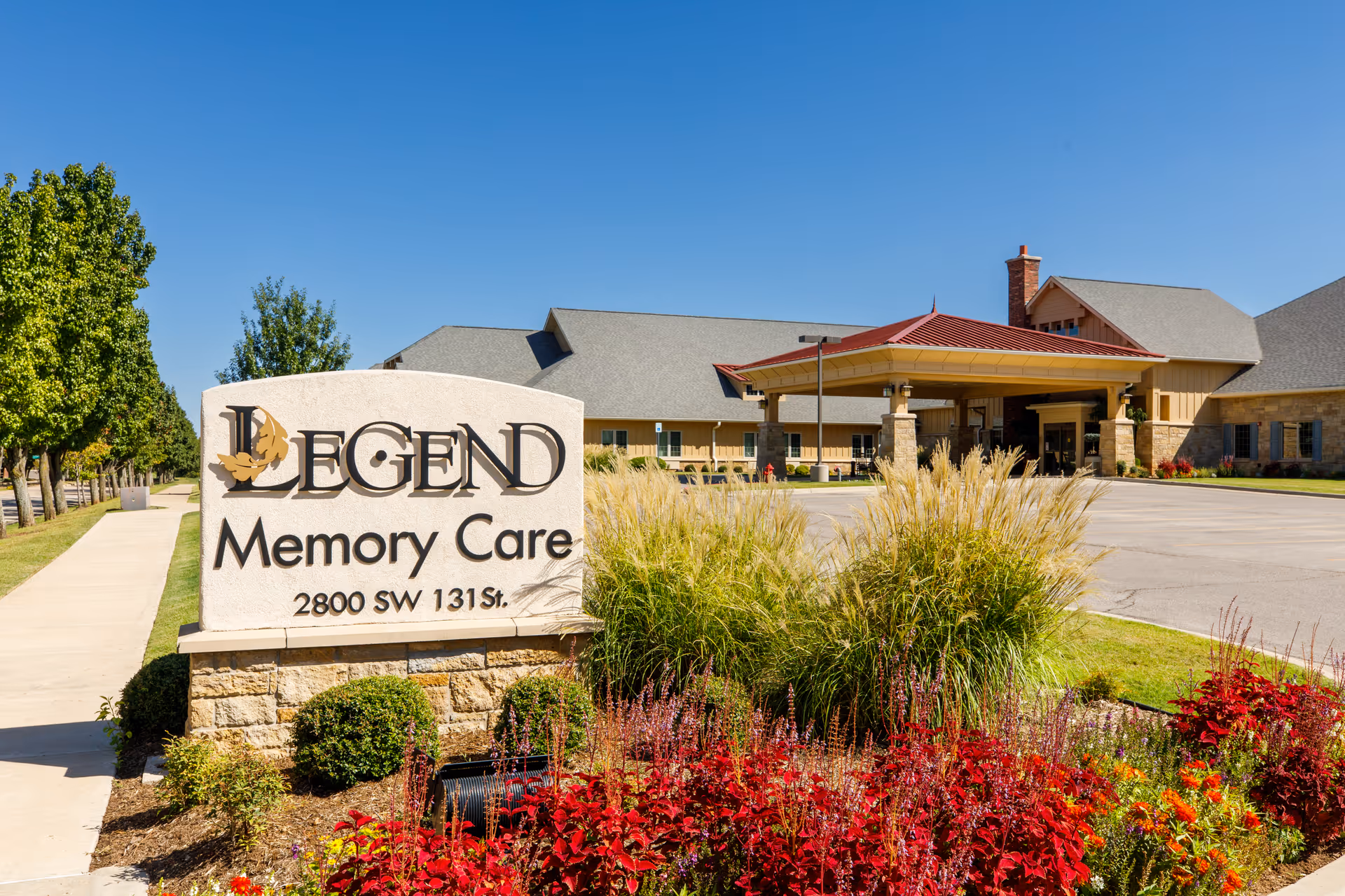 Stone sign reading 'Legend Memory Care' in front of the facility's landscaped entrance and driveway under a clear blue sky.