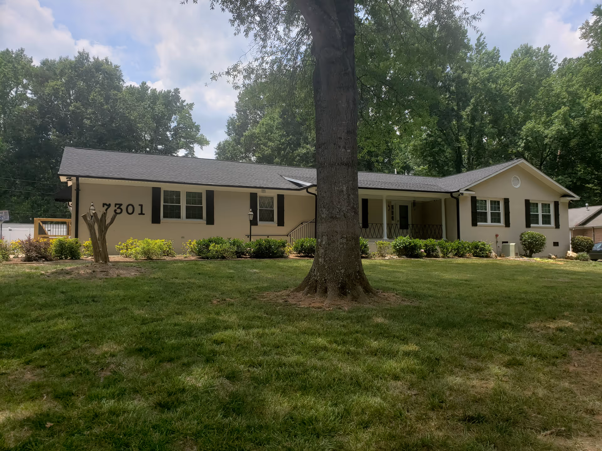Single-story residential building with beige exterior walls, black shutters, and a dark roof, surrounded by green grass and trees under a partly cloudy sky. The building has the number 7301 displayed on the front wall and a covered porch area with railings.