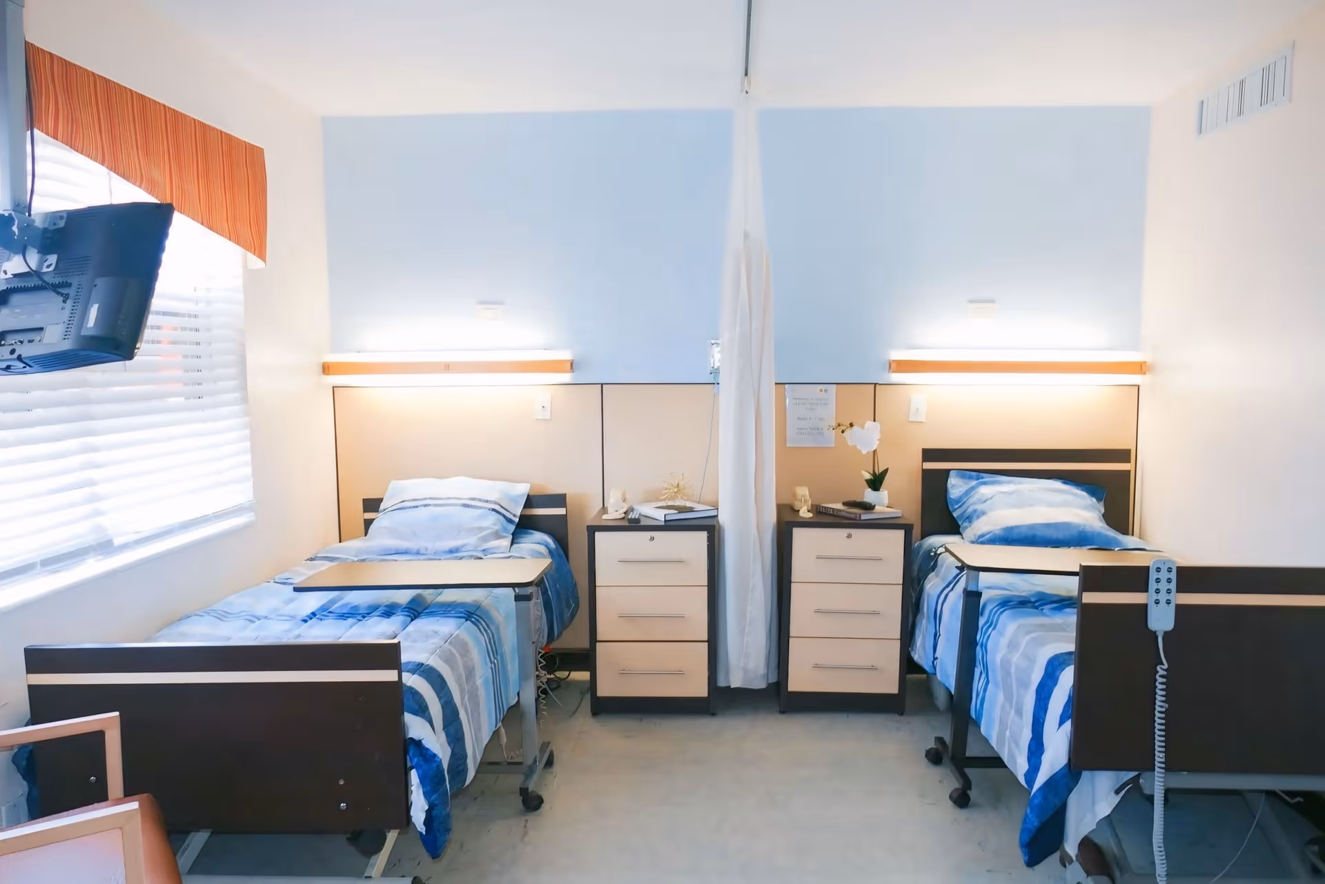 A hospital-style room with two single beds separated by a white privacy curtain. Each bed has blue and white striped bedding, an overbed table, and a bedside cabinet with drawers. There is a window with blinds and an orange valance on the left side, and a wall-mounted TV near the window.